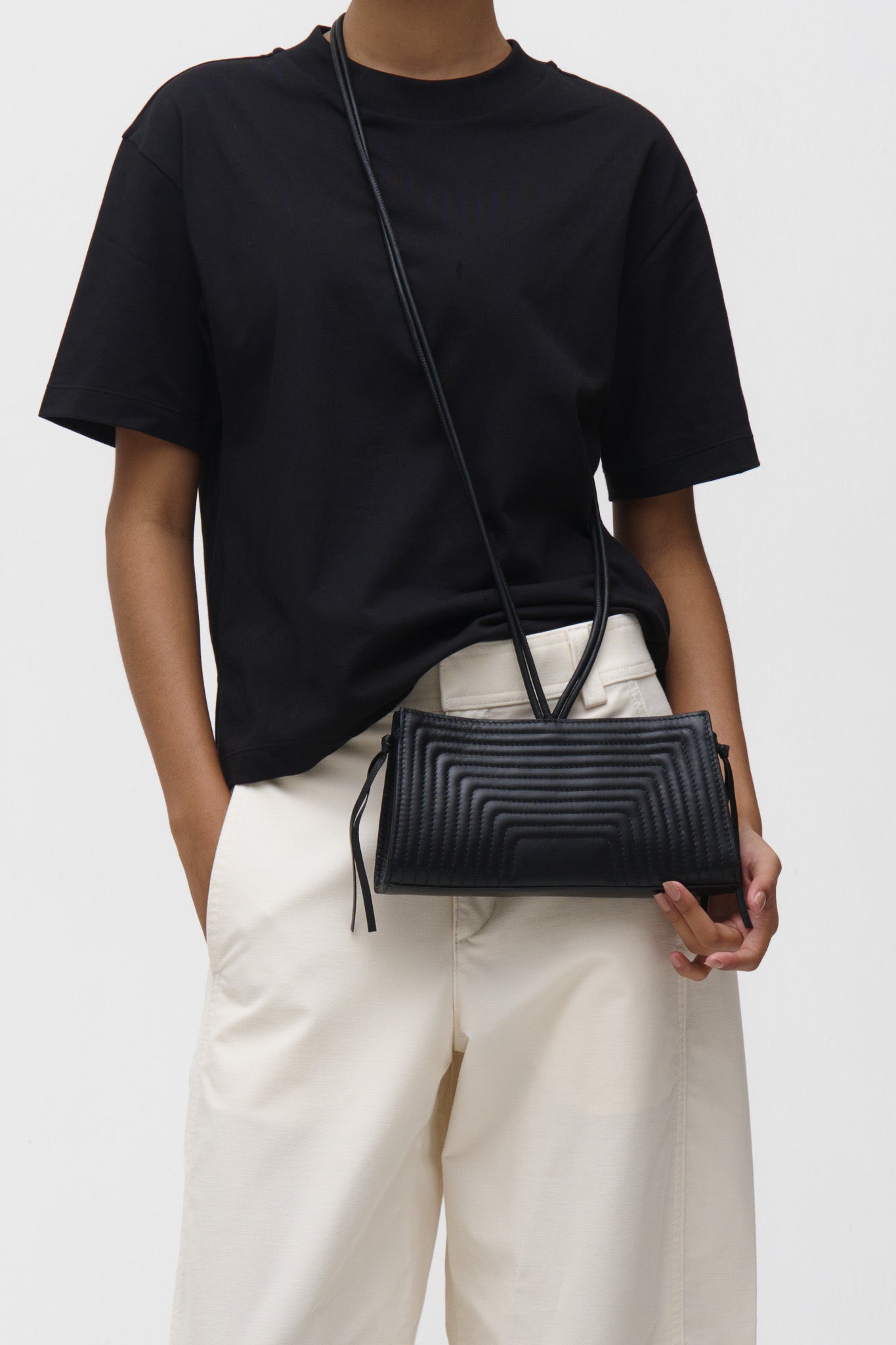 Person wearing a black t-shirt and light-colored pants holding a black crossbody bag against a white background