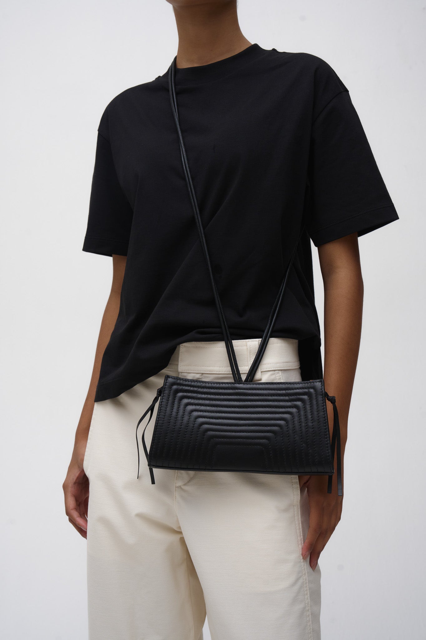 Person wearing a black t-shirt and light-colored pants with a black crossbody bag on a white background